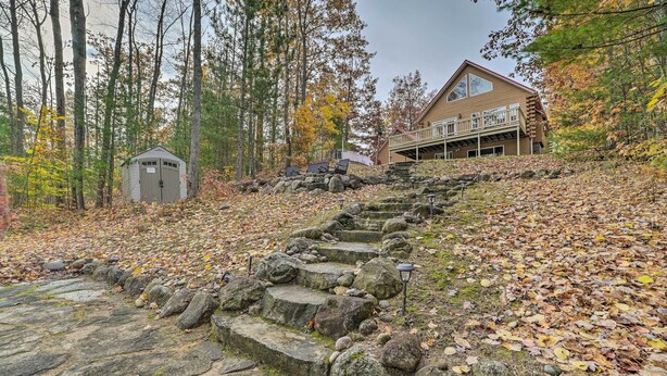 A cabin with a stone staircase leading up to a wooden deck, surrounded by autumn foliage and tall trees.