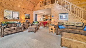 A cozy cabin interior with a wooden staircase, leather sofas, a dining area, and a view through a window.
