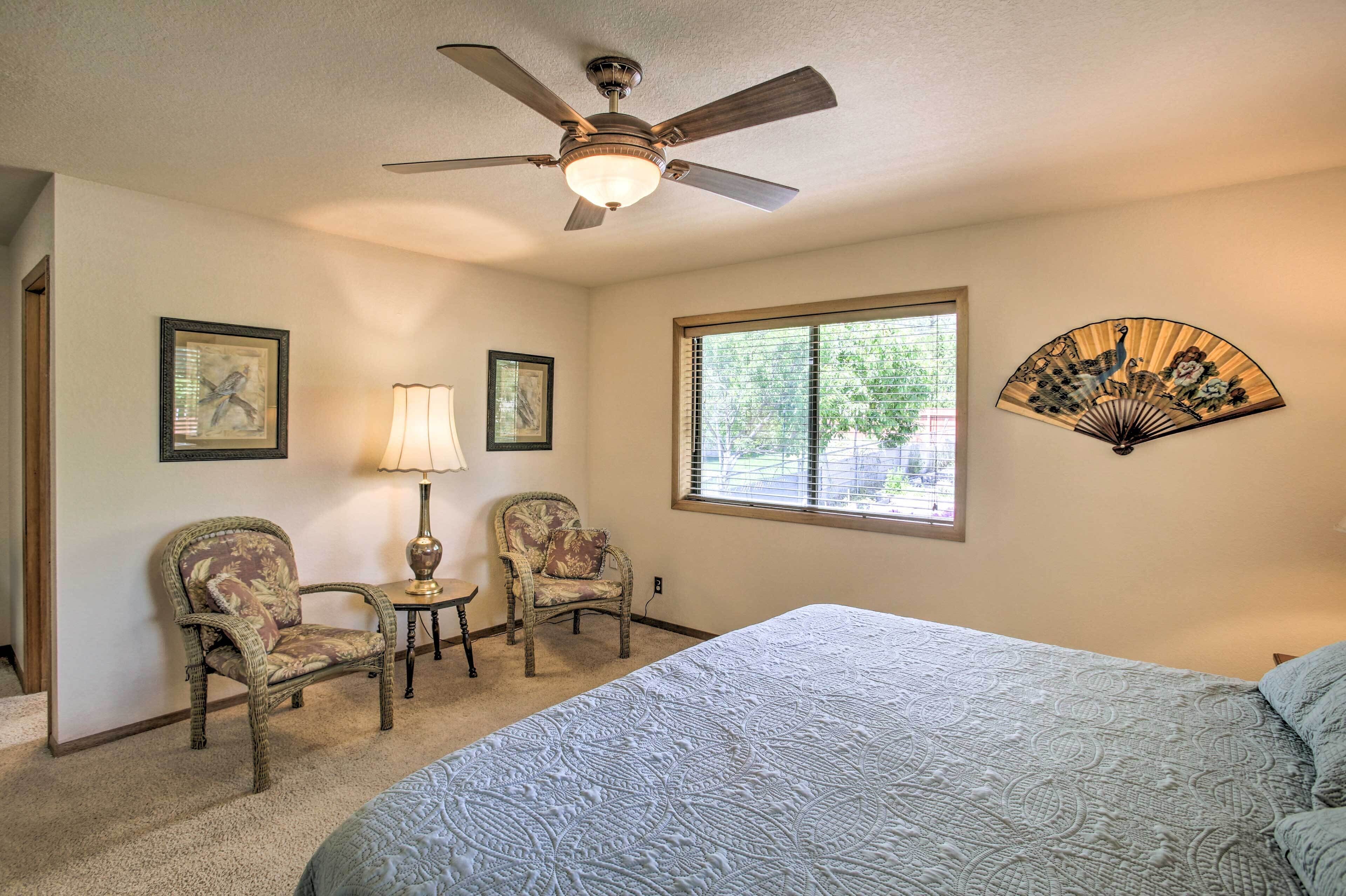 A bedroom with a bed, two chairs, a table, a fan, and a window with blinds.