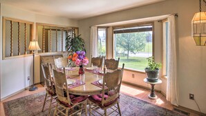 A dining area with a round table, wooden chairs, a vase with flowers, and a view of the outdoors through large windows.