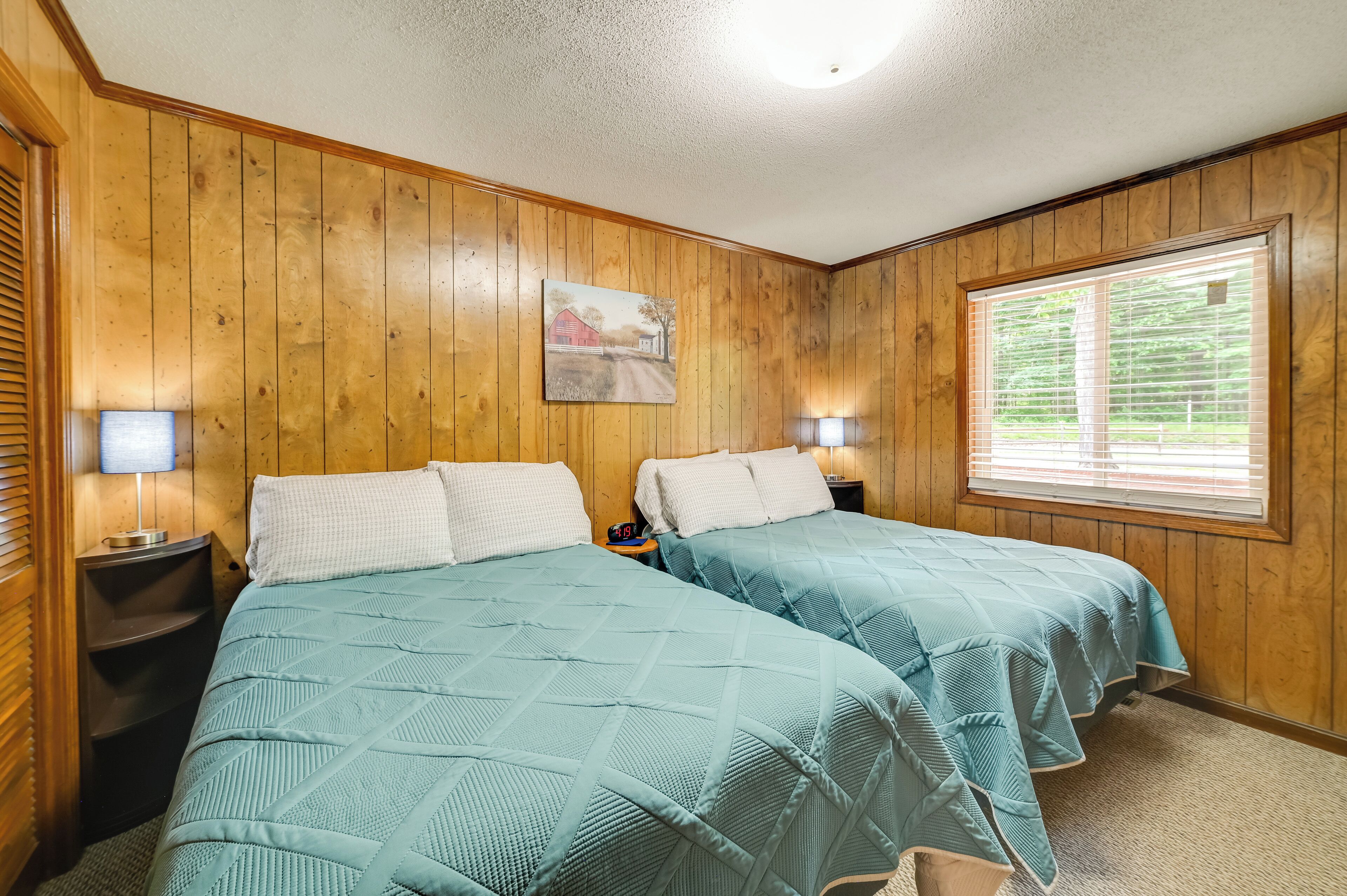 Une pièce aux murs recouverts de lambris en bois, deux lits avec une literie de couleur turquoise, un tableau accroché au mur et une fenêtre avec des stores.
