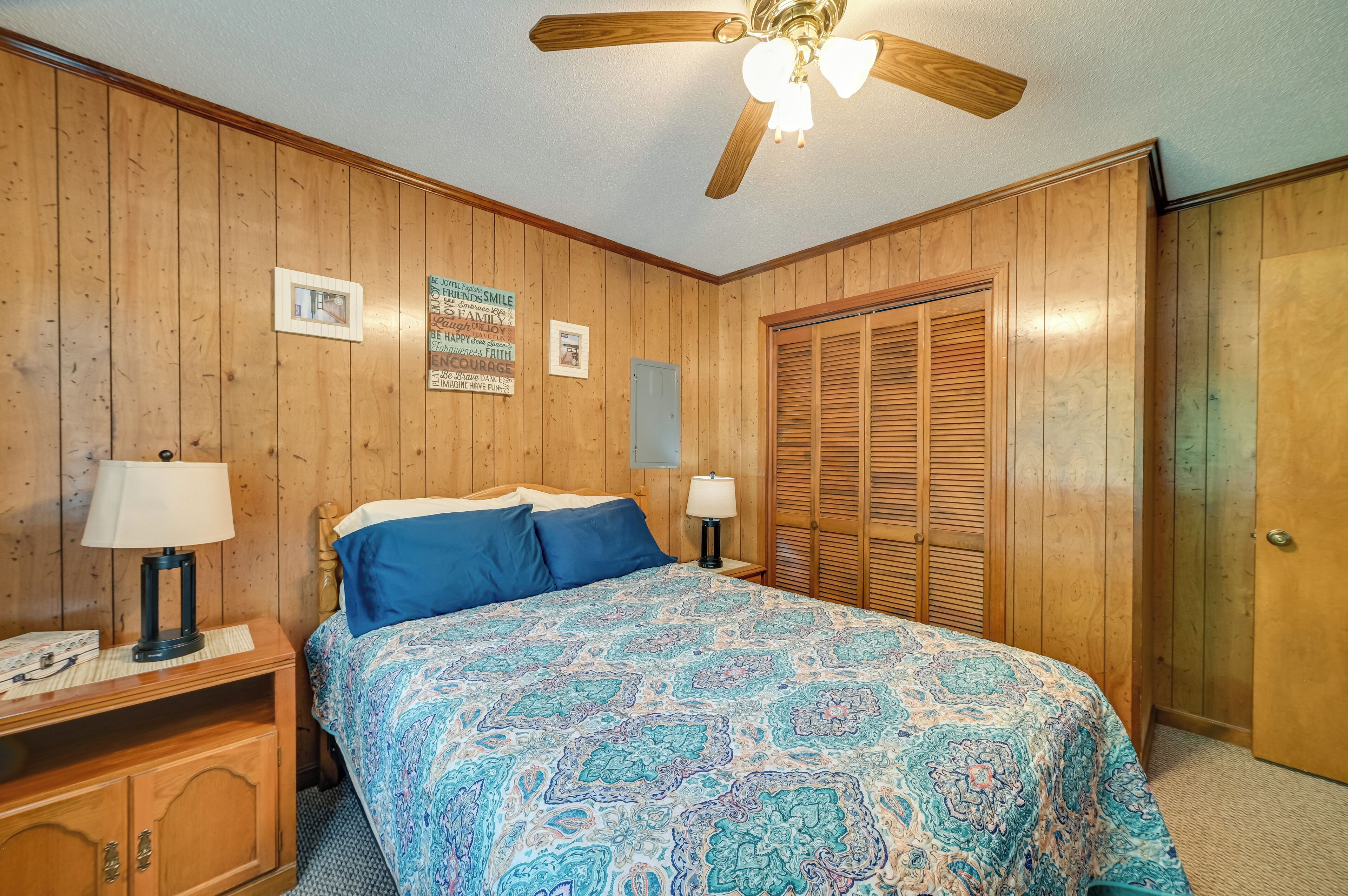 Quarto com painéis de madeira, uma cama com colcha estampada, duas mesas de cabeceira com abajures, um ventilador de teto e um guarda-roupa de madeira.