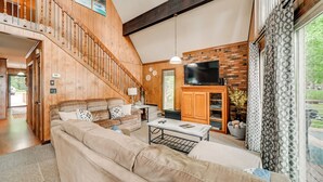 A spacious living room with a high ceiling, wooden staircase, a flat-screen TV, and a large window with patterned curtains. - Cozy Dover Home w/ Boat Dock, Fire Pit & Deck! (Dover)