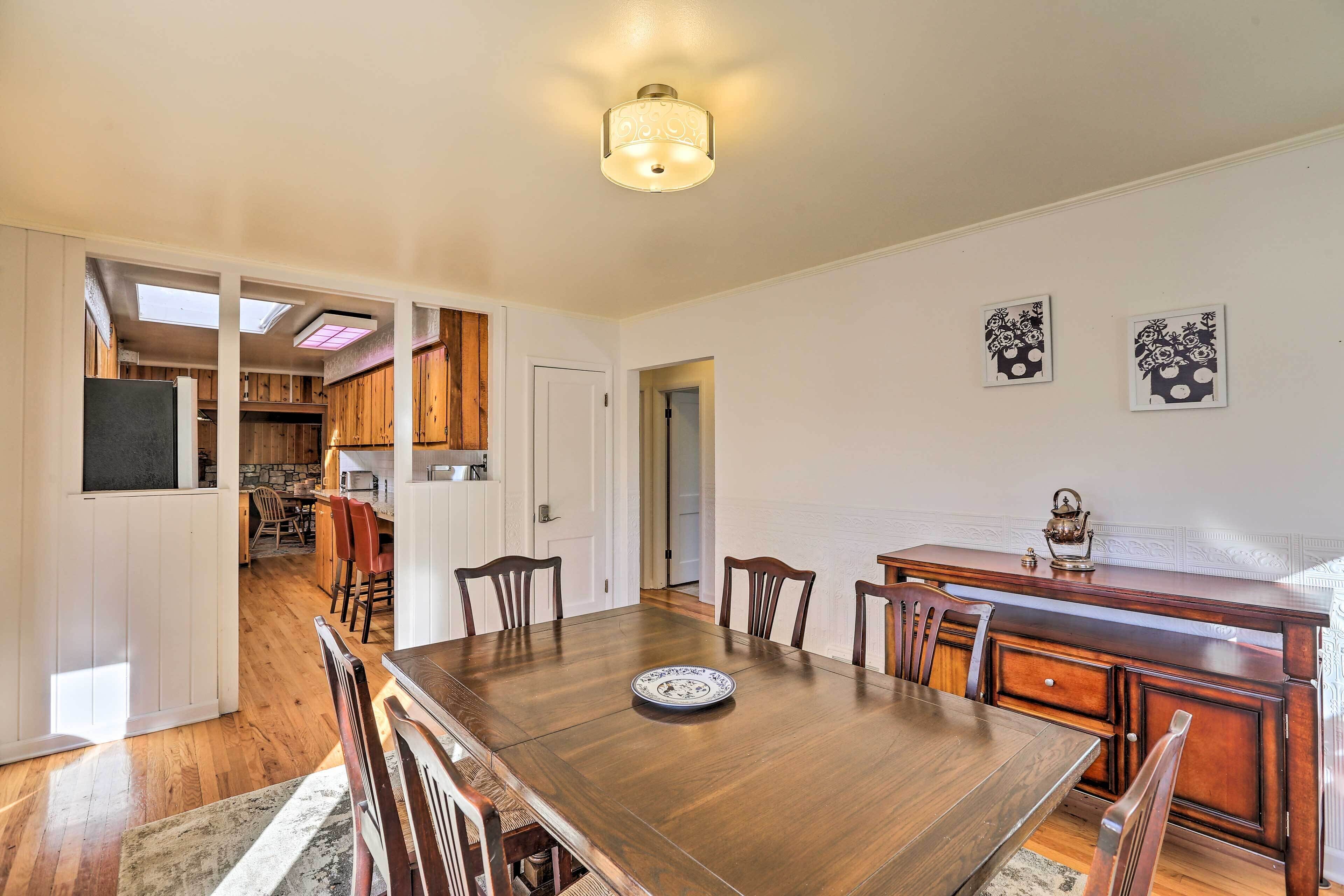 A dining area with a wooden table, chairs, and a decorative plate. There is a kitchen in the background with wooden cabinets and a window.