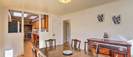 A dining area with a wooden table, chairs, and a decorative plate. There is a kitchen in the background with wooden cabinets and a window.