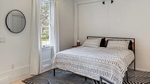 A bedroom with a bed, a window with curtains, a round mirror, and a patterned rug. - Steps to NYC Train! Charming Carriage House Loft (Glen Ridge)