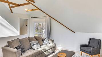 A modern living room with a sofa, armchair, and coffee table, wooden beams on the ceiling, and a window with curtains.