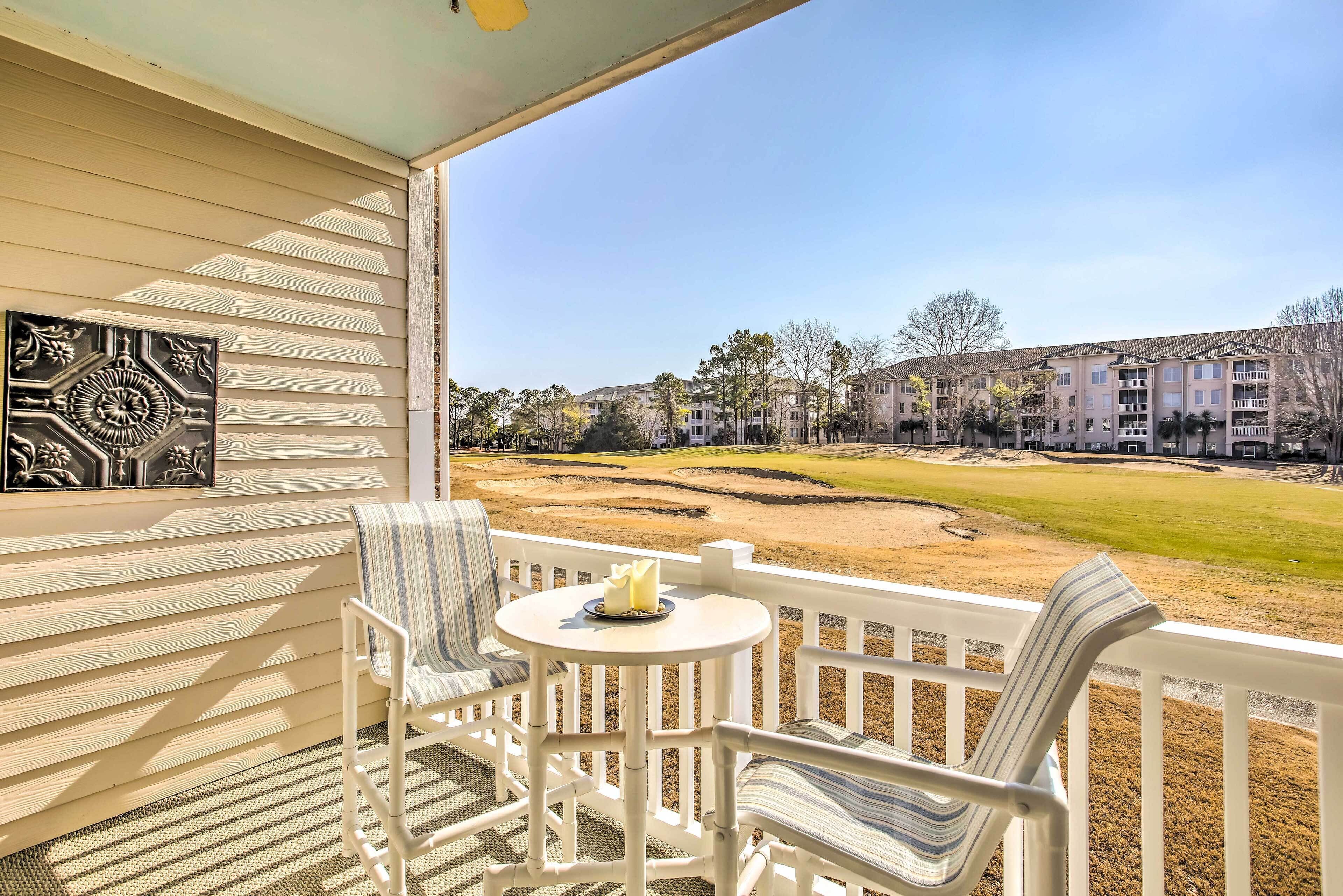 A balcony with white wicker chairs and a small table, overlooking a golf course and residential buildings.