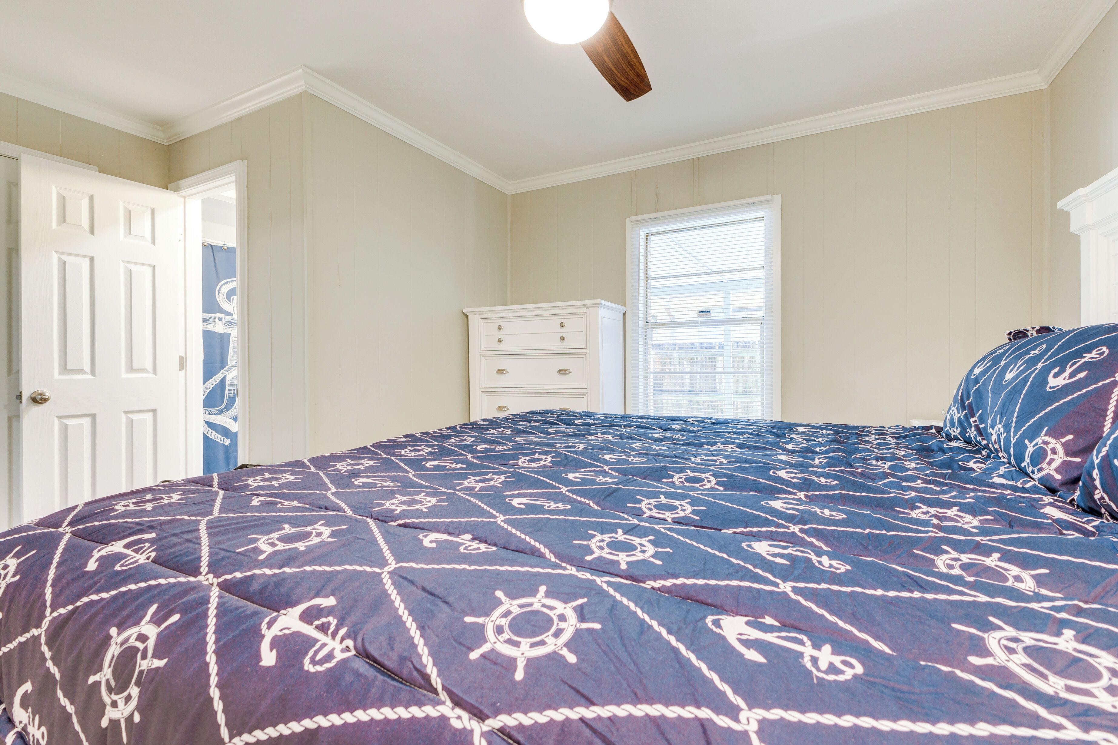 A bedroom with a navy blue bedspread featuring nautical designs, a white dresser, and a window with blinds.