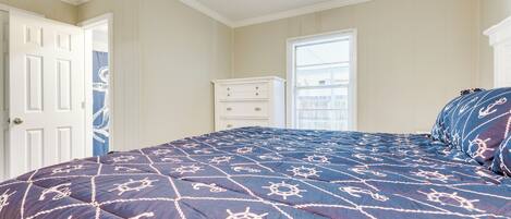 A bedroom with a navy blue bedspread featuring nautical designs, a white dresser, and a window with blinds.