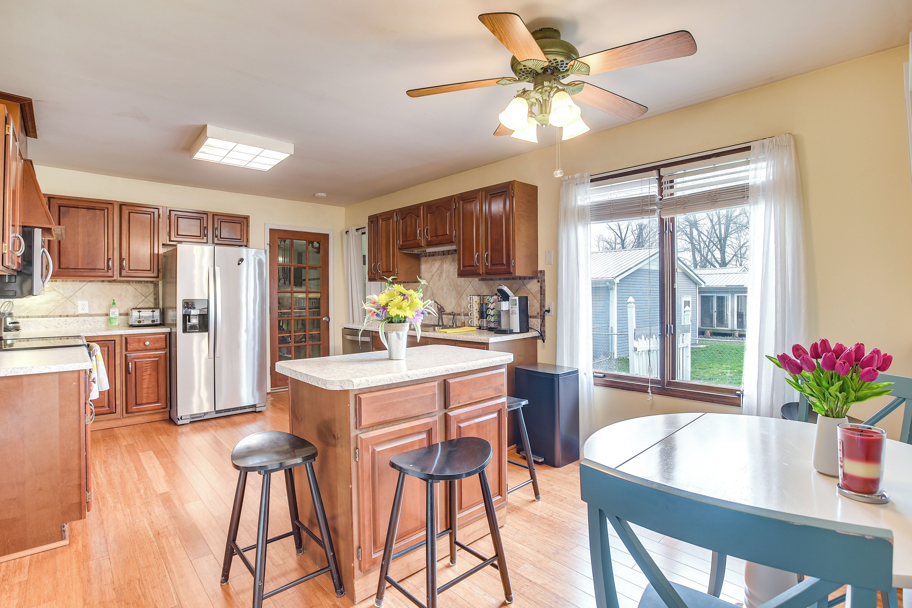 A kitchen with wooden cabinets, a stainless steel refrigerator, a ceiling fan, and a window with a view of a house.