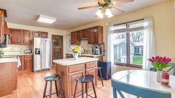 A kitchen with wooden cabinets, a stainless steel refrigerator, a ceiling fan, and a window with a view of a house.