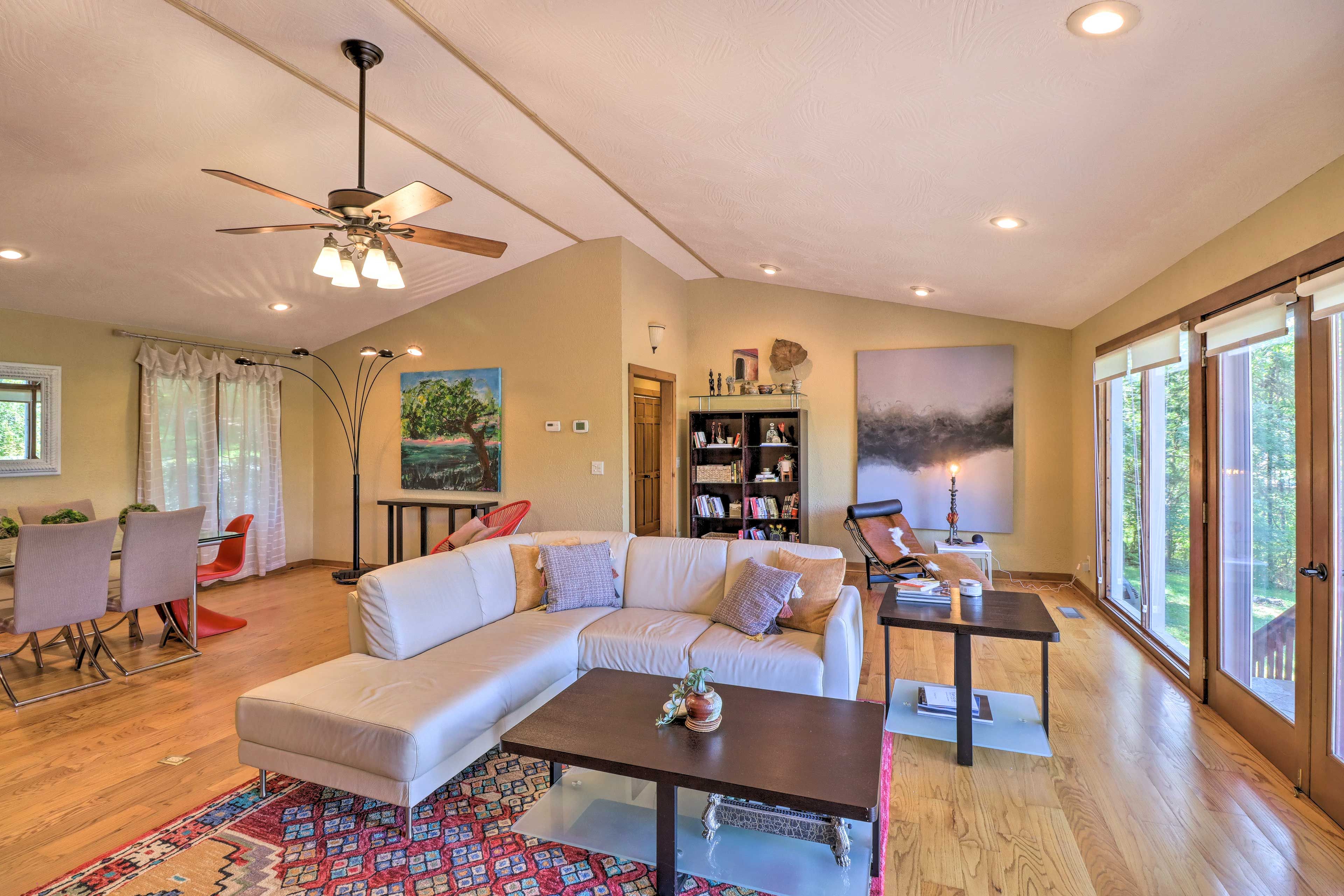 A spacious living room with a sectional sofa, a dining area with chairs, a ceiling fan, and large windows offering a view of the outdoors.