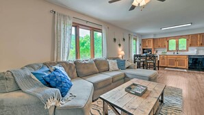 A spacious living room with a large sectional sofa, a coffee table, and a ceiling fan. Adjacent to the living room is a kitchen with wooden cabinets and a large window. - Pet-friendly Waynesville Home w/ Mountain Views! (Waynesville)