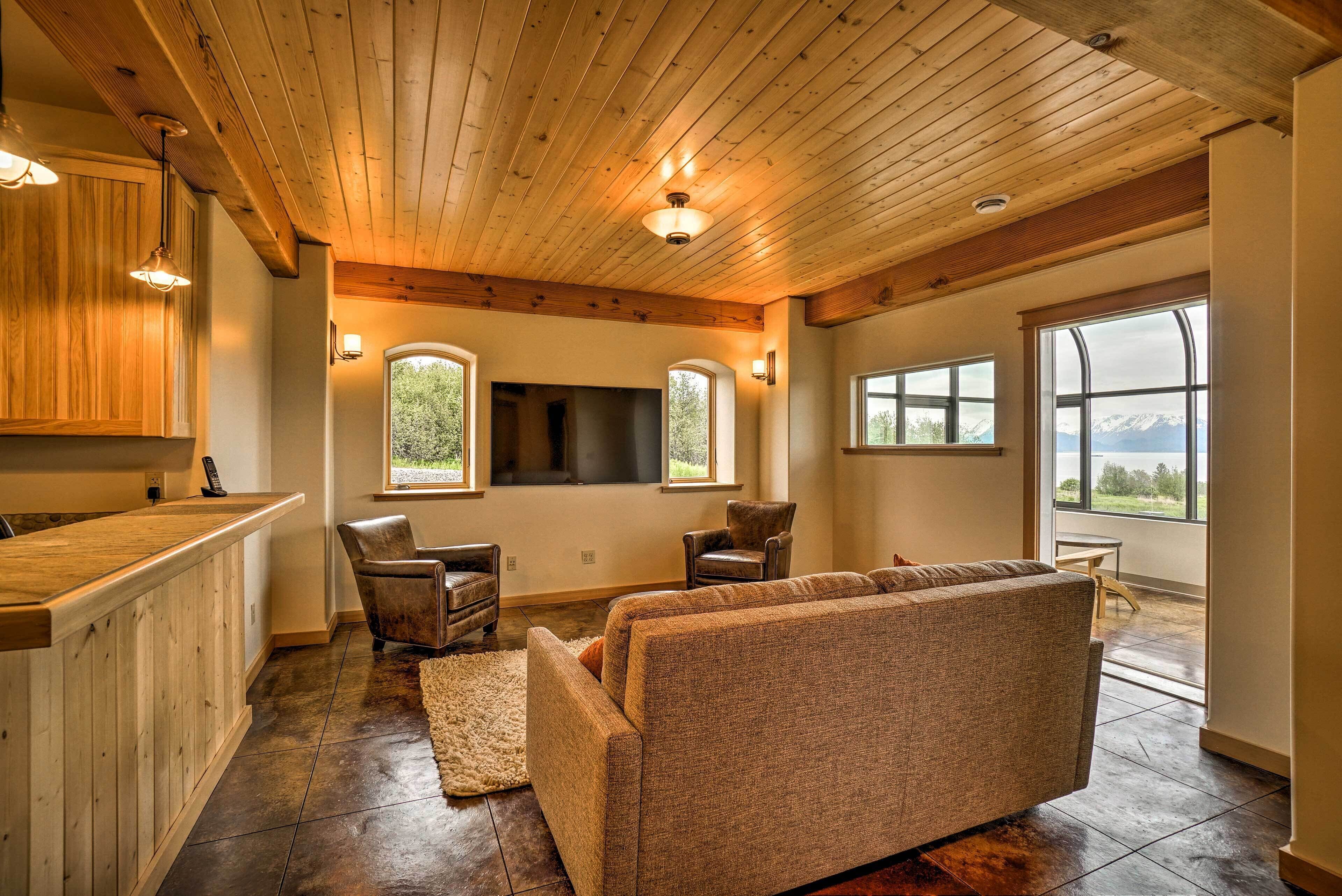 A spacious living area with a wooden ceiling, a sofa, two armchairs, a fireplace, and large windows offering mountain views.