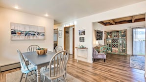 Un coin repas avec une table et des chaises en bois, un tableau accroché au mur, et une vue sur la cuisine et le salon par une porte ouverte.