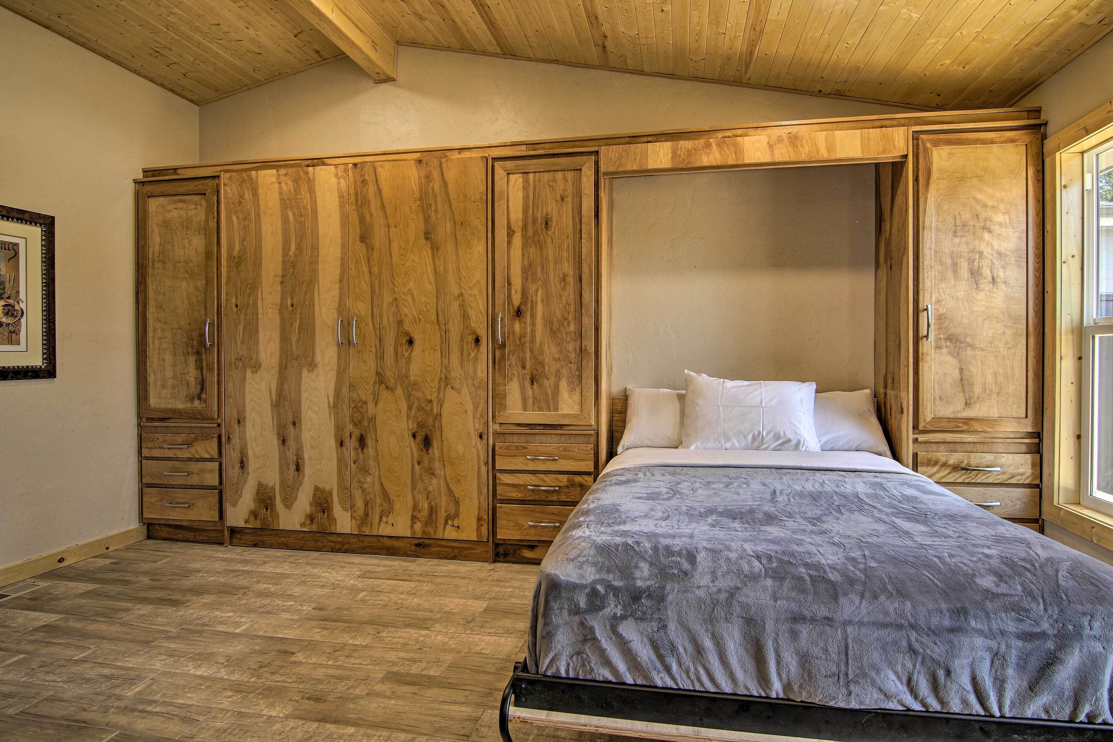 Une chambre avec une grande armoire en bois, un lit recouvert d’une couette grise et une fenêtre sur le côté droit.