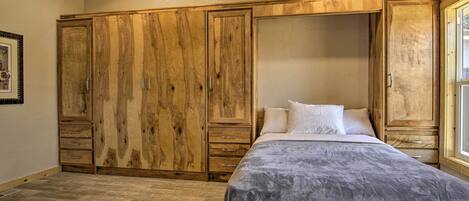 Une chambre avec une grande armoire en bois, un lit recouvert d’une couette grise et une fenêtre sur le côté droit.