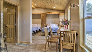 A room with a wooden ceiling, a bed with white bedding, a dining table with chairs, and a window with a view of greenery. - Mountain Cabin, Walk to Dining/memorial Park! (Woodland Park)
