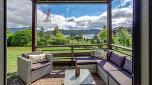 A covered outdoor seating area with wicker furniture and cushions, overlooking a scenic landscape with mountains and a lake.