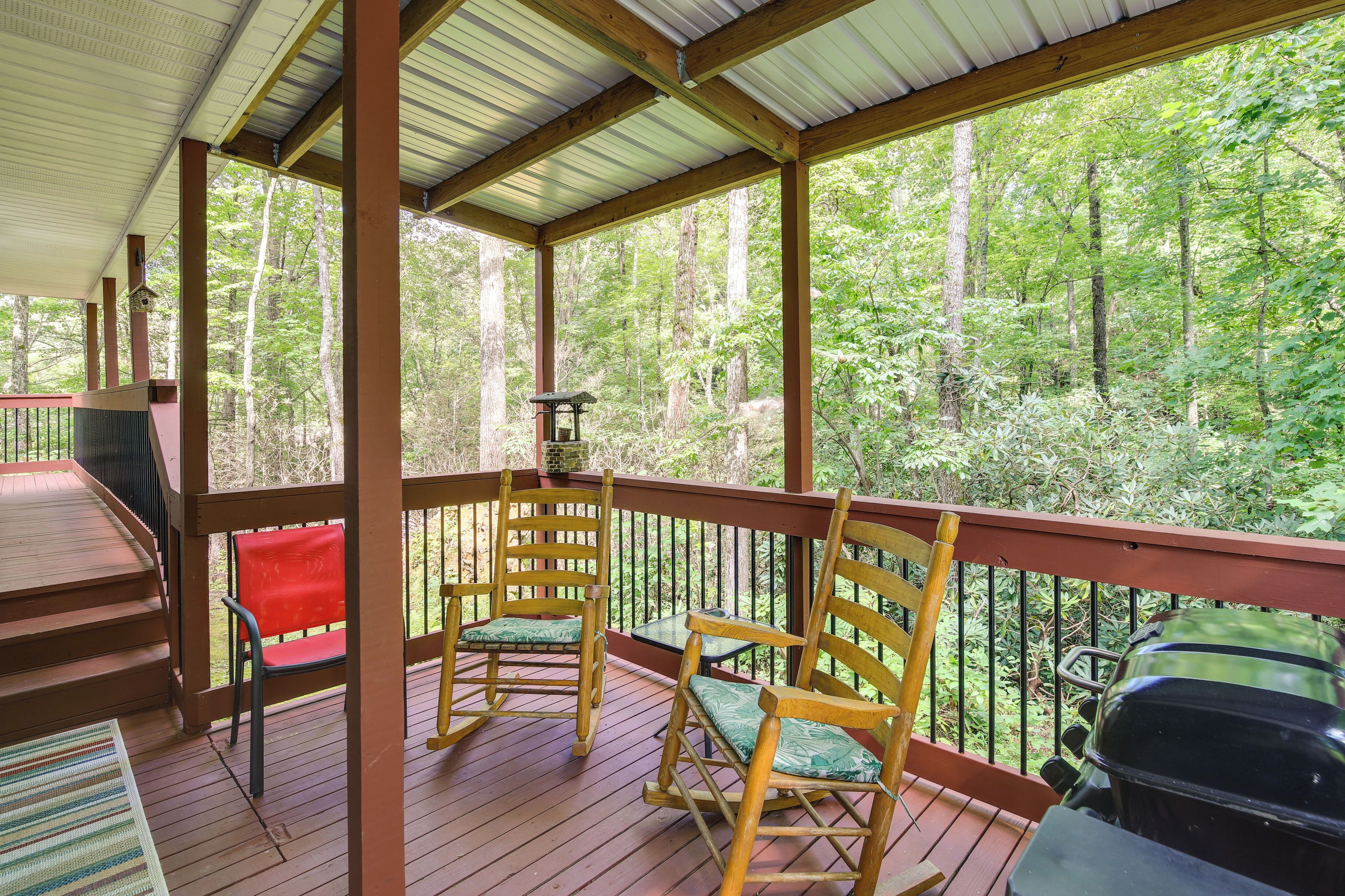 A covered outdoor deck with wooden chairs and a table, overlooking a forested area.