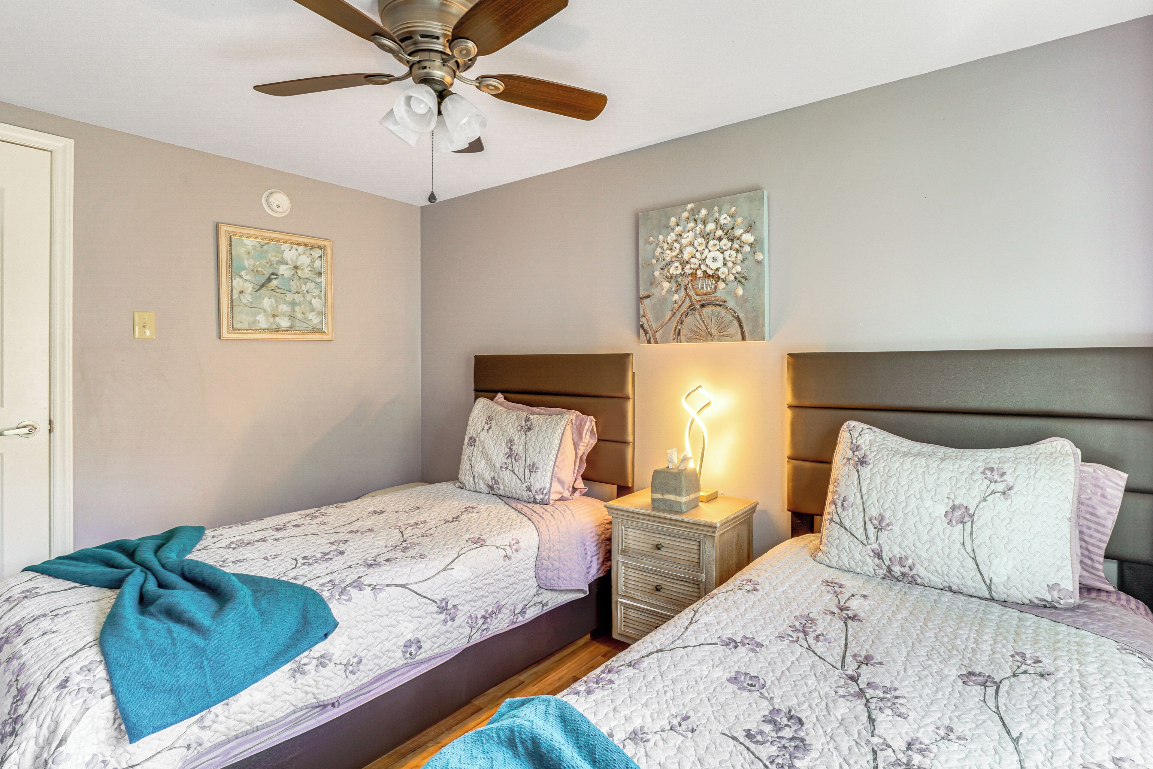 A bedroom with two beds, a ceiling fan, a nightstand with a lamp, and wall art.