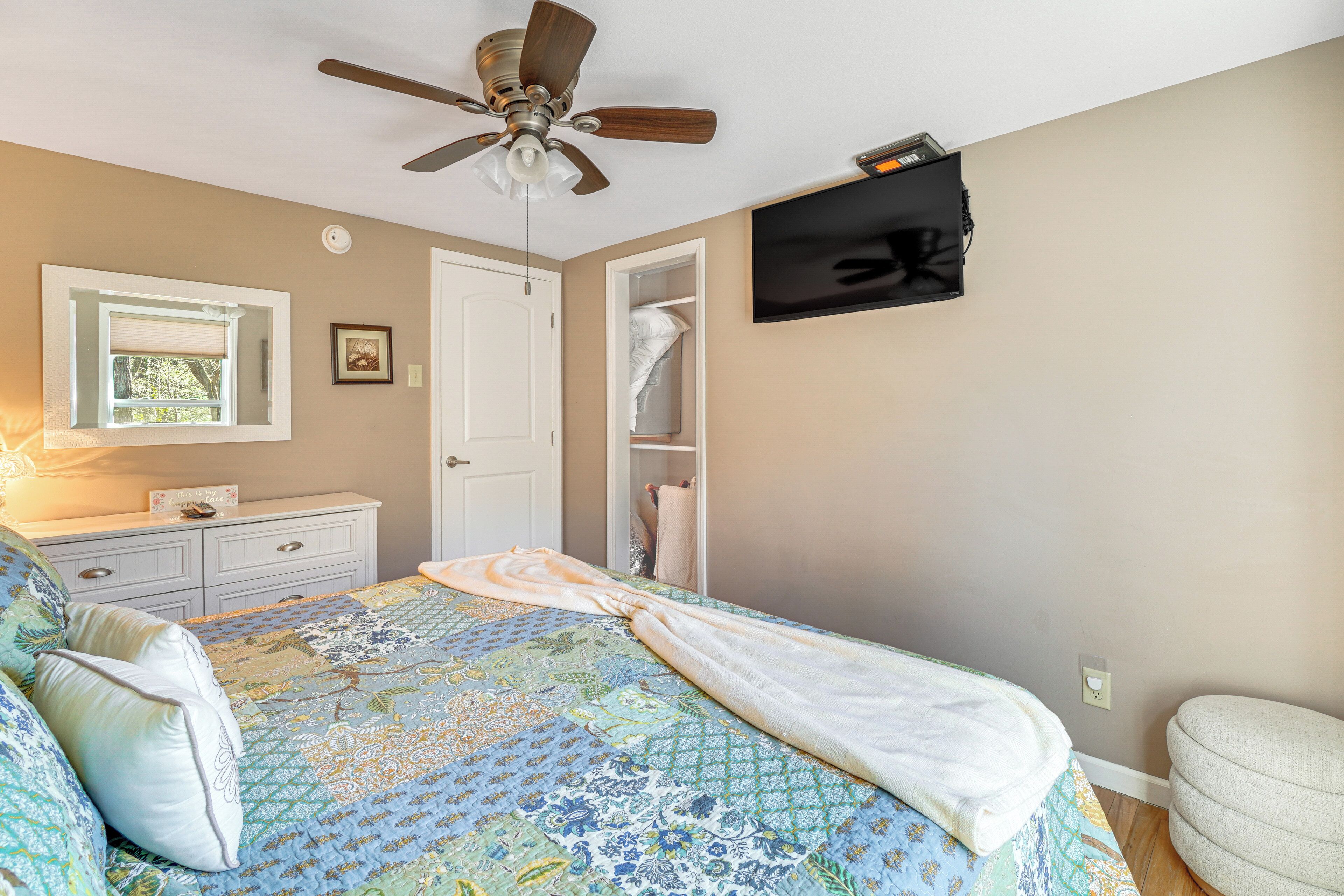 A bedroom with a bed, a ceiling fan, a TV mounted on the wall, a dresser, and a closet.