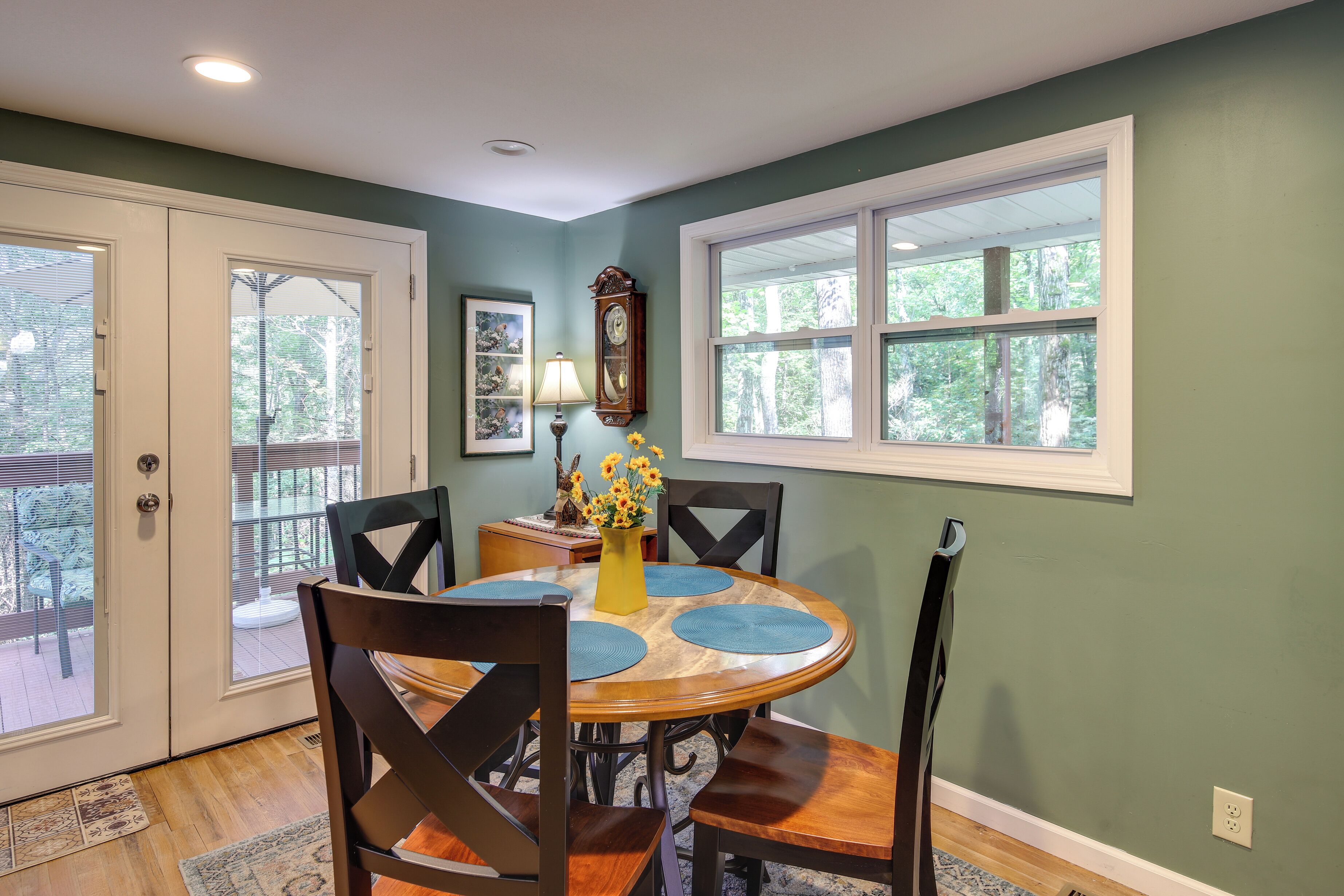 A dining area with a round table, four chairs, a vase with flowers, a clock, and a window with a view of trees.