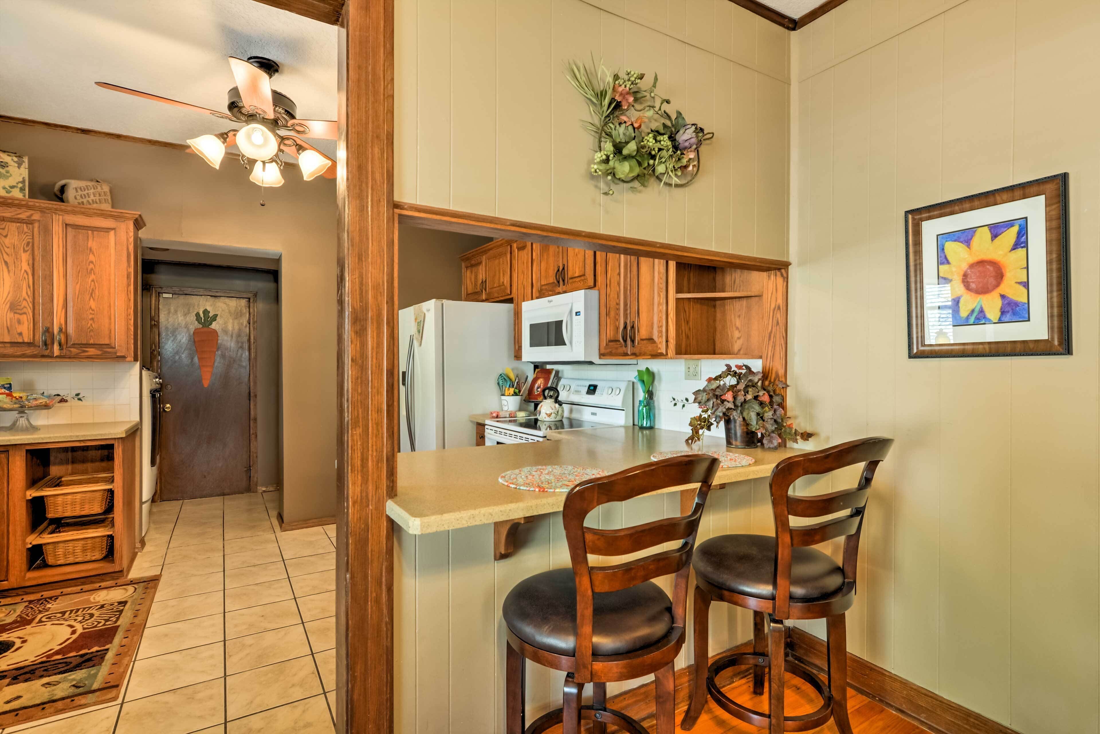 A kitchen with wooden cabinets, a refrigerator, a microwave, and a sink. There are two wooden chairs at the kitchen island.
