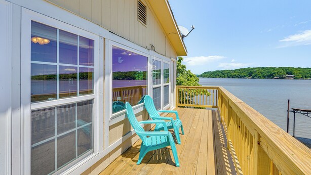A wooden deck with two turquoise Adirondack chairs, large windows, and a view of a lake and hills. - Deck w/ Covered Dock! Lakefront Ozark Cottage (Climax Springs)