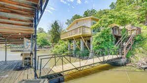 A wooden deck with a bridge leading to a house with a balcony, surrounded by greenery and a body of water. - Deck w/ Covered Dock! Lakefront Ozark Cottage (Climax Springs)