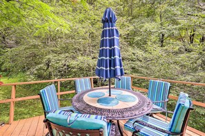 A round table with a blue umbrella, surrounded by striped chairs on a wooden deck with a view of dense greenery. - Furnished Deck + Home Gym: Bakersville Retreat! (Bakersville)