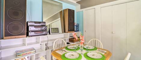 A dining area with a wooden table set for four, surrounded by white chairs. A shelf above the table holds various items including a JVC speaker, a stereo system, and a Monopoly game box.