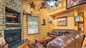A log cabin interior with a stone fireplace, leather sofa, and a ceiling fan. - Porch & Fire Pit: Rustic Log Cabin in Smokies! (Cosby)