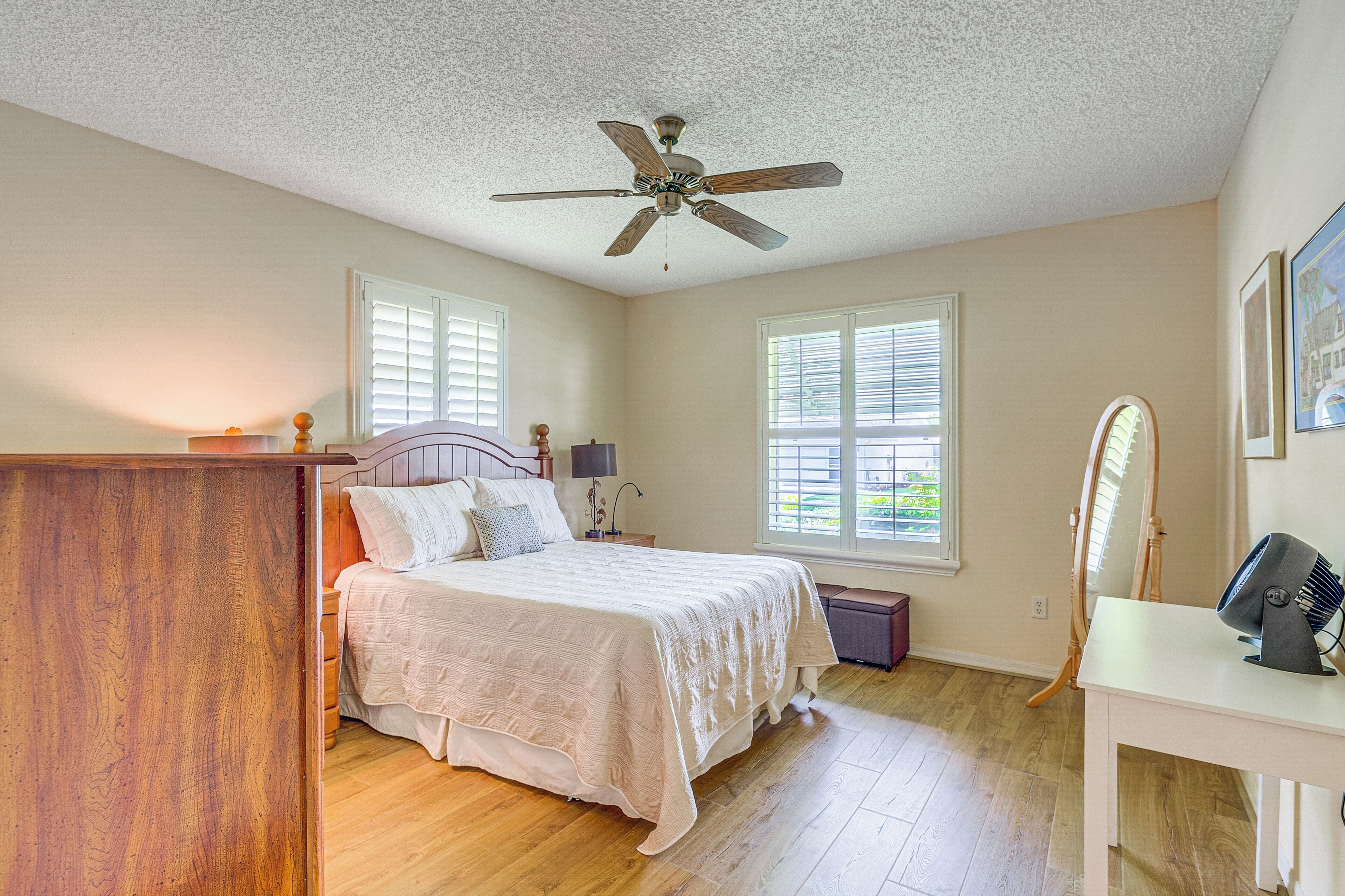 A bedroom with a bed, a ceiling fan, a dresser, a vanity with a mirror, and two windows with blinds.