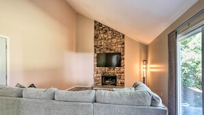 A living room with a stone fireplace, a large gray sectional sofa, and a large window providing a view of greenery outside.