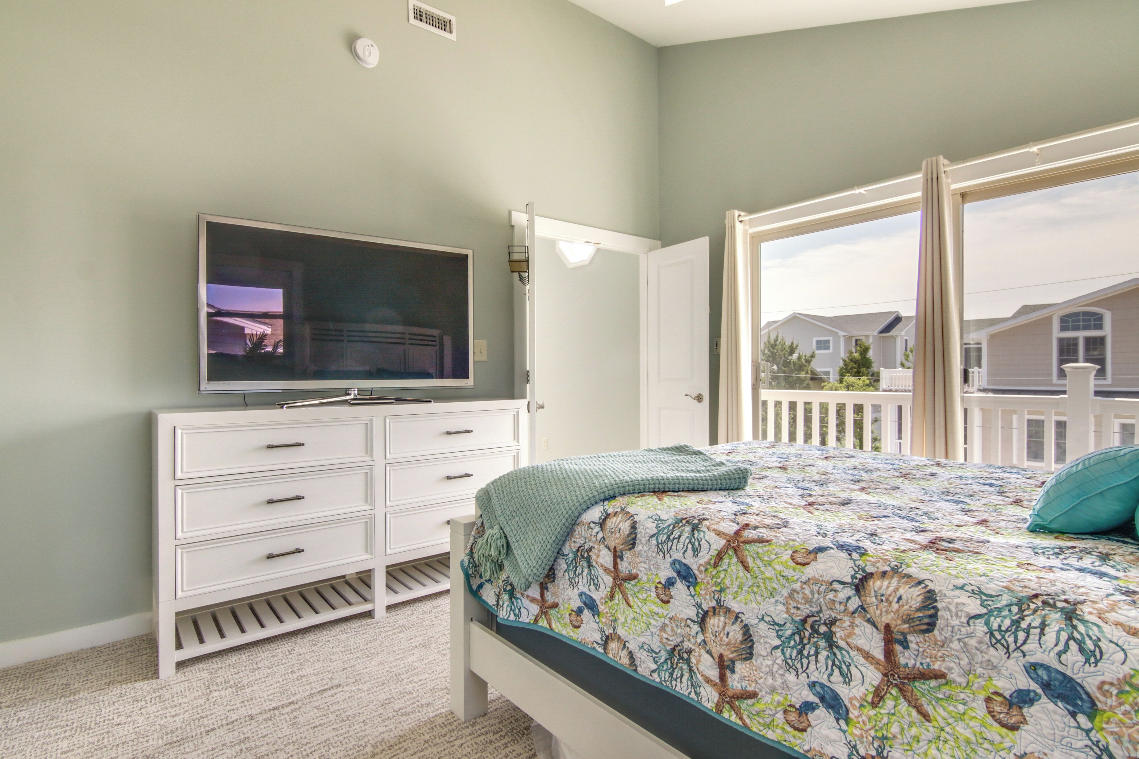 A bedroom with a bed, a dresser, a television, and a window with a view of houses.