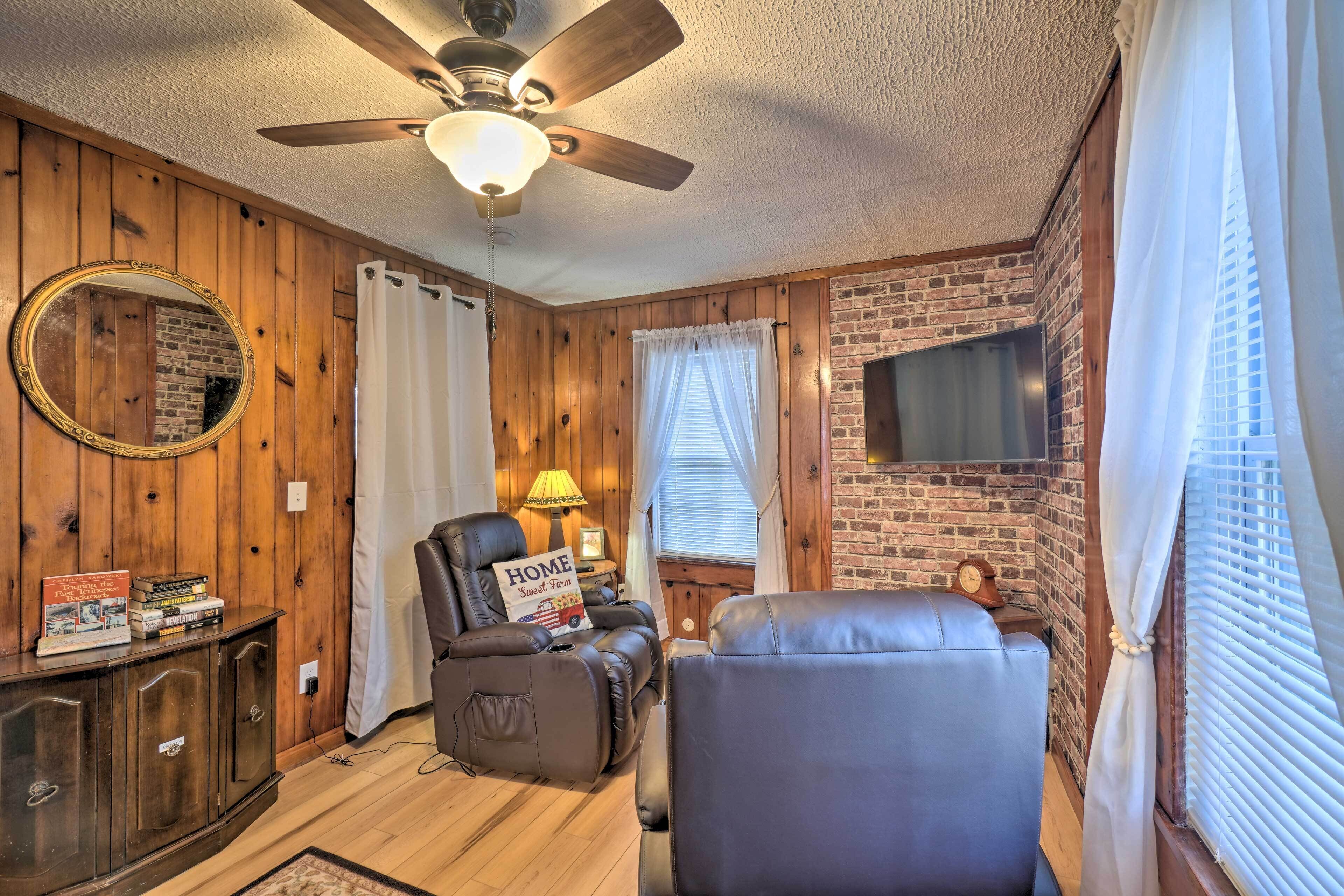A cozy room with a ceiling fan, a leather recliner, a massage chair, a TV mounted on a brick wall, and a wooden cabinet with books.