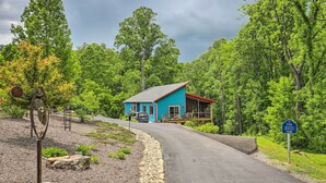 Une maison bleue avec un toit rouge et une terrasse, entourée d’une végétation luxuriante et bordée d’un chemin de gravier.