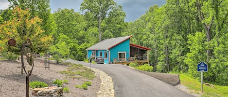 A blue house with a red roof and a deck, surrounded by lush greenery and a gravel path.
