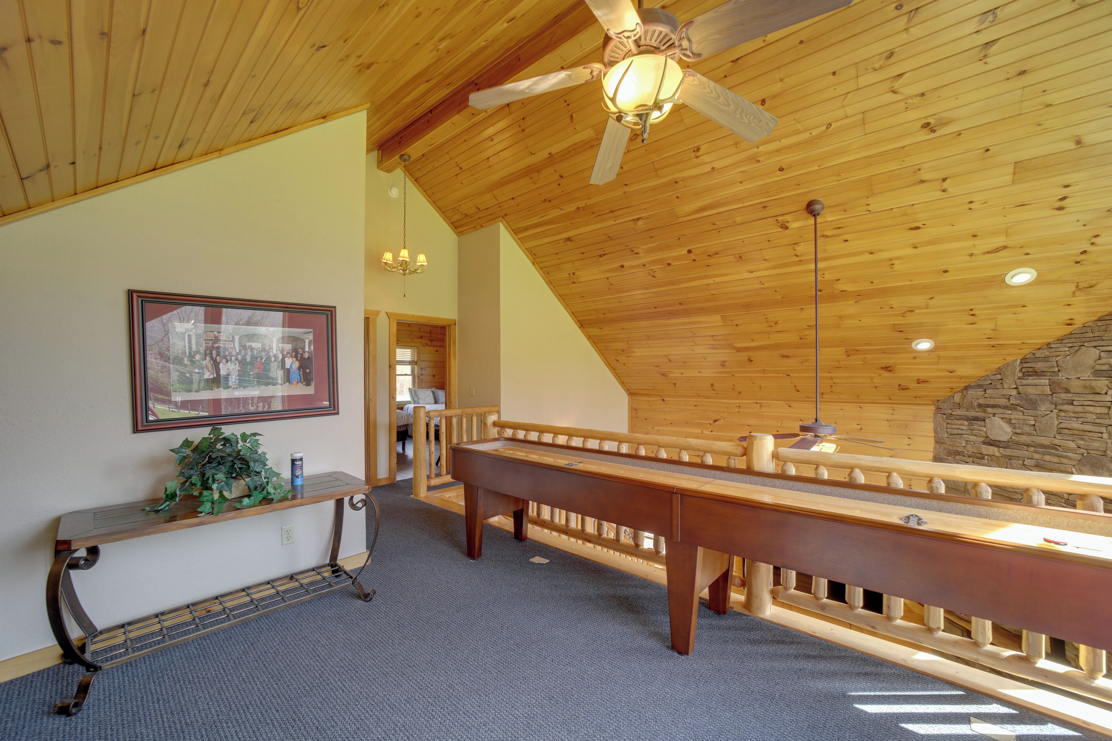 A spacious room with a high ceiling, wooden beams, a sloped roof, a stone wall, a wooden bench, a small table with a plant, and a framed picture on the wall.