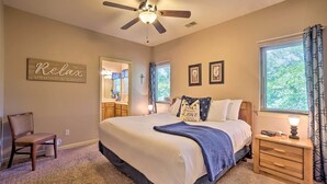 A bedroom with a bed, a chair, a nightstand, a ceiling fan, a window with curtains, and a wall with a sign.
