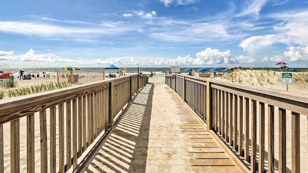 Une promenade en bois mène à une plage où se trouvent des parasols et un panneau indiquant qu’une opération de restauration des dunes est en cours.