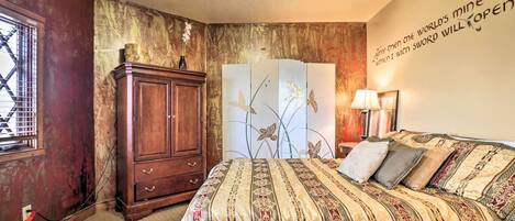 Un dormitorio con cama, ventilador de techo, armario de madera, pared decorativa con una cita y ventana con barrotes.
