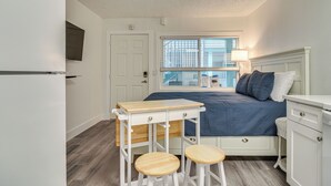 A compact bedroom with a bed, a small table with stools, a white dresser, and a window with blinds.