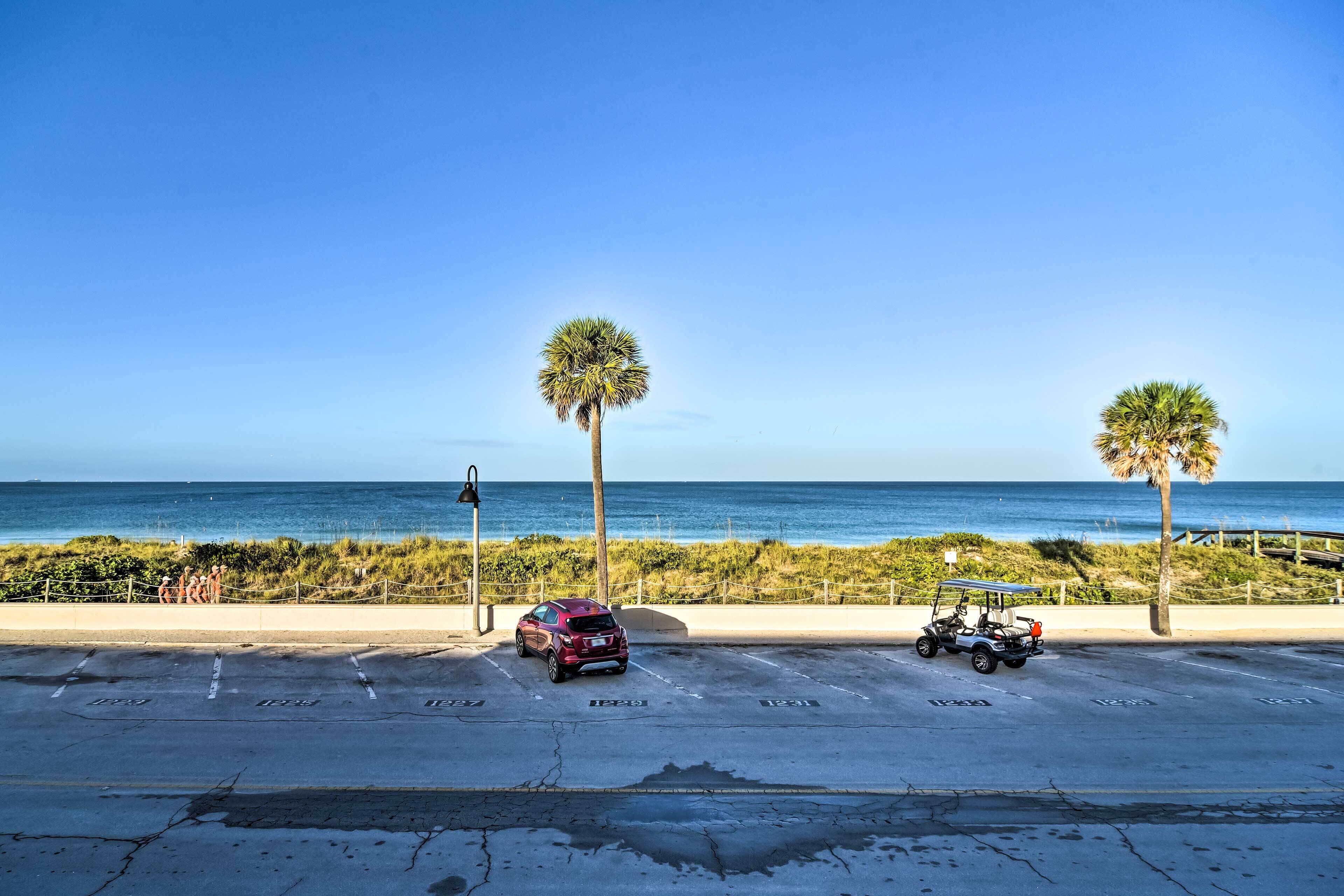 A parking lot with a red SUV and a golf cart, palm trees, and a view of the ocean.