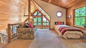 Ein gemütliches Zimmer mit Holzbalkendecke, einem Bett mit gemustertem Überwurf, einer Couch mit Kissen und einem großen Fenster mit Blick auf Bäume.