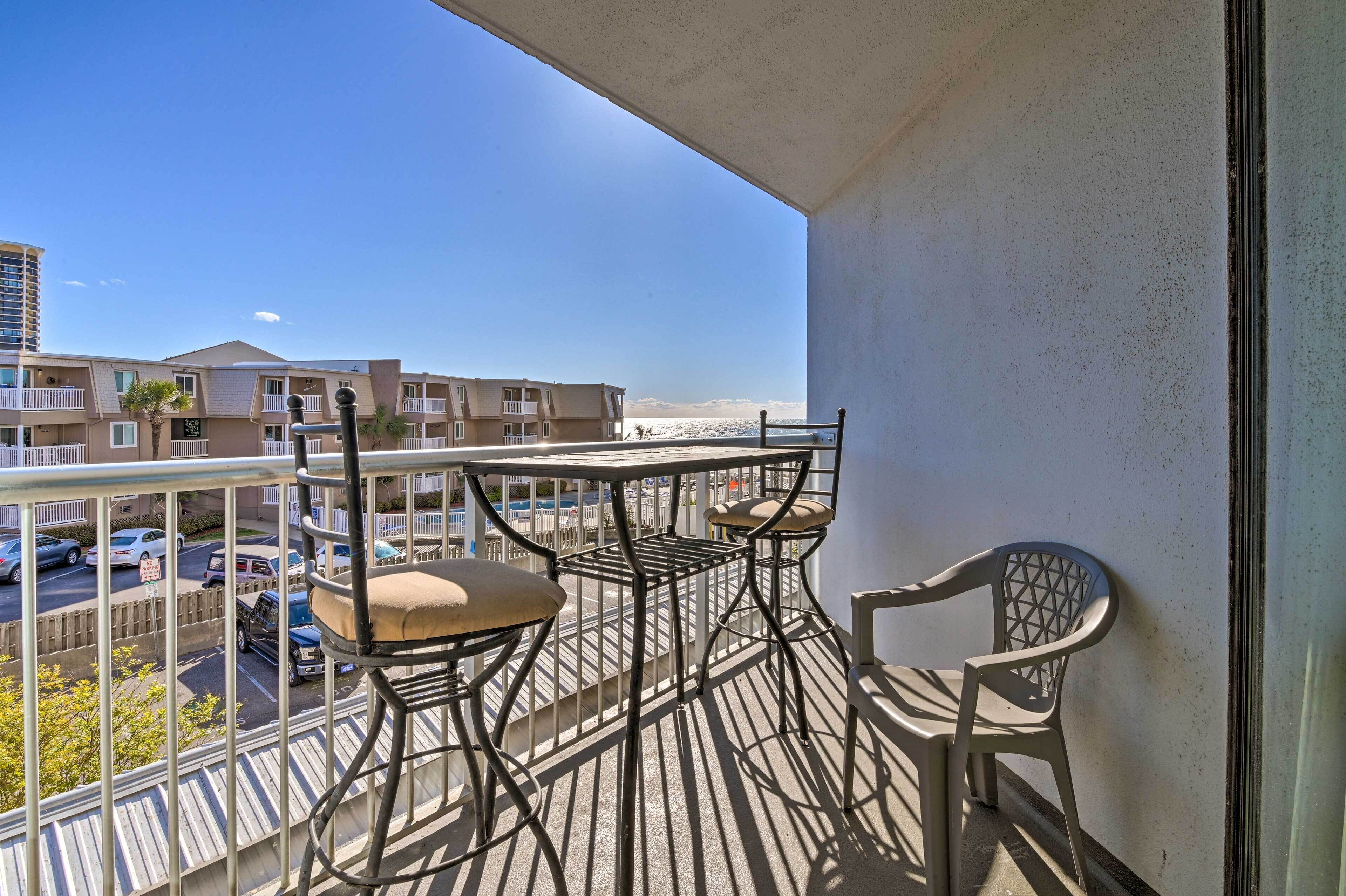 A balcony with a table and chairs, overlooking a street with parked cars and buildings.