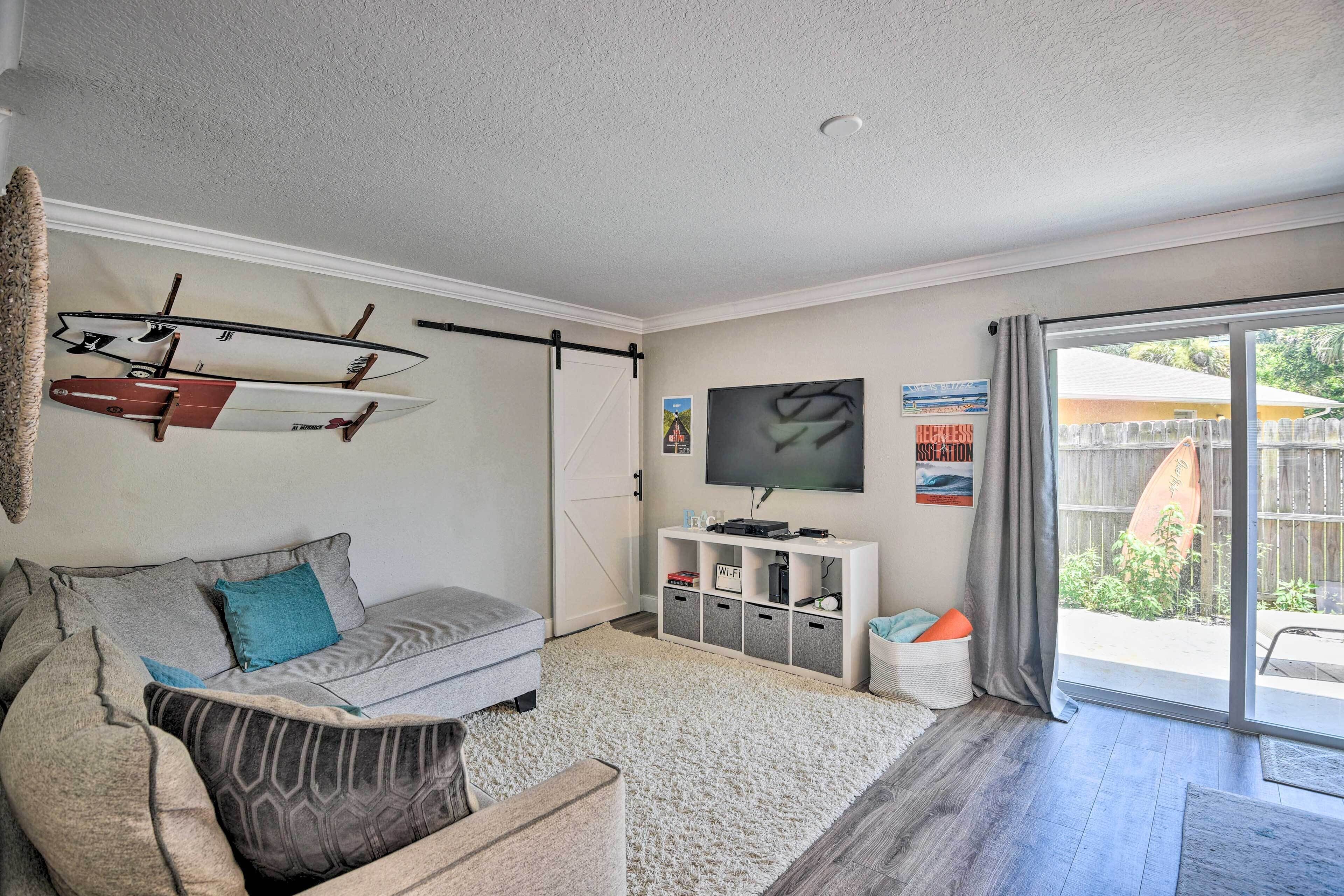 A living room with a grey sofa, a flat-screen TV, a white entertainment unit, and a sliding glass door to the outdoors.