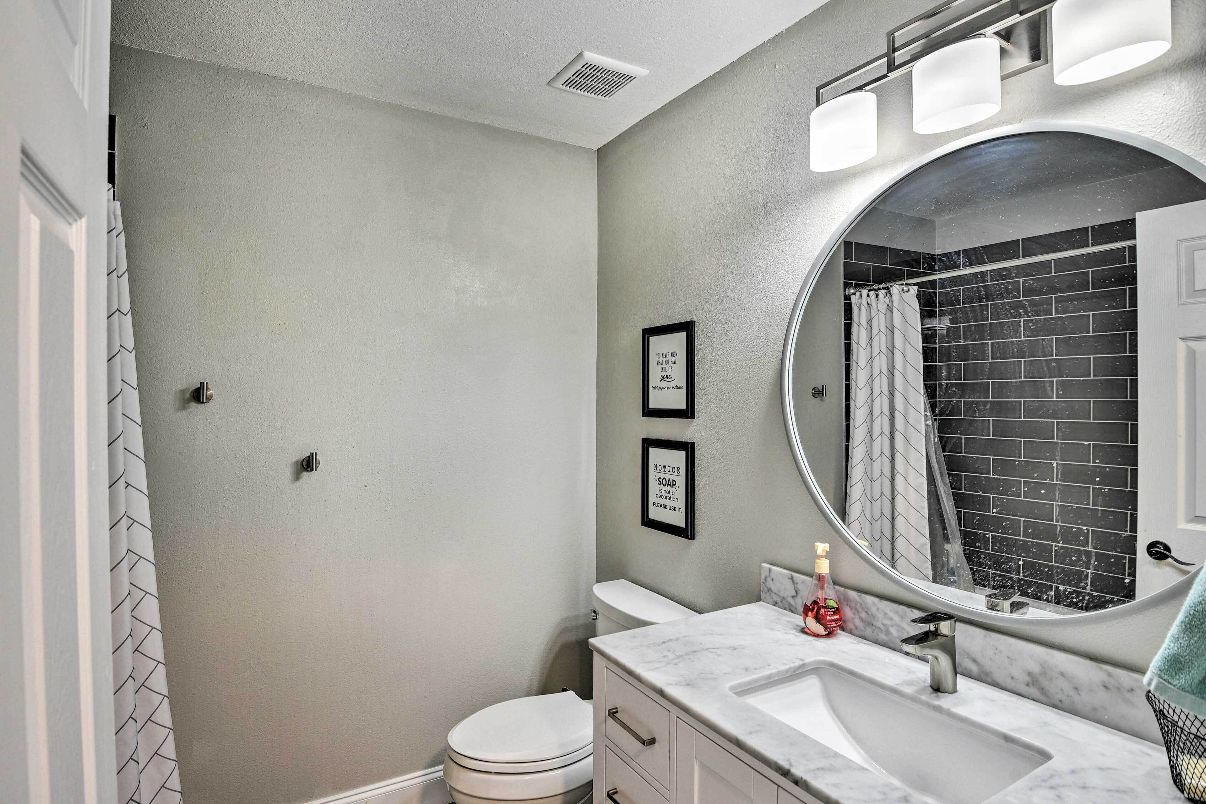 Une salle de bain avec des toilettes blanches, un lavabo doté d’un plan de travail en marbre, un miroir rond et une douche aux carreaux noirs.